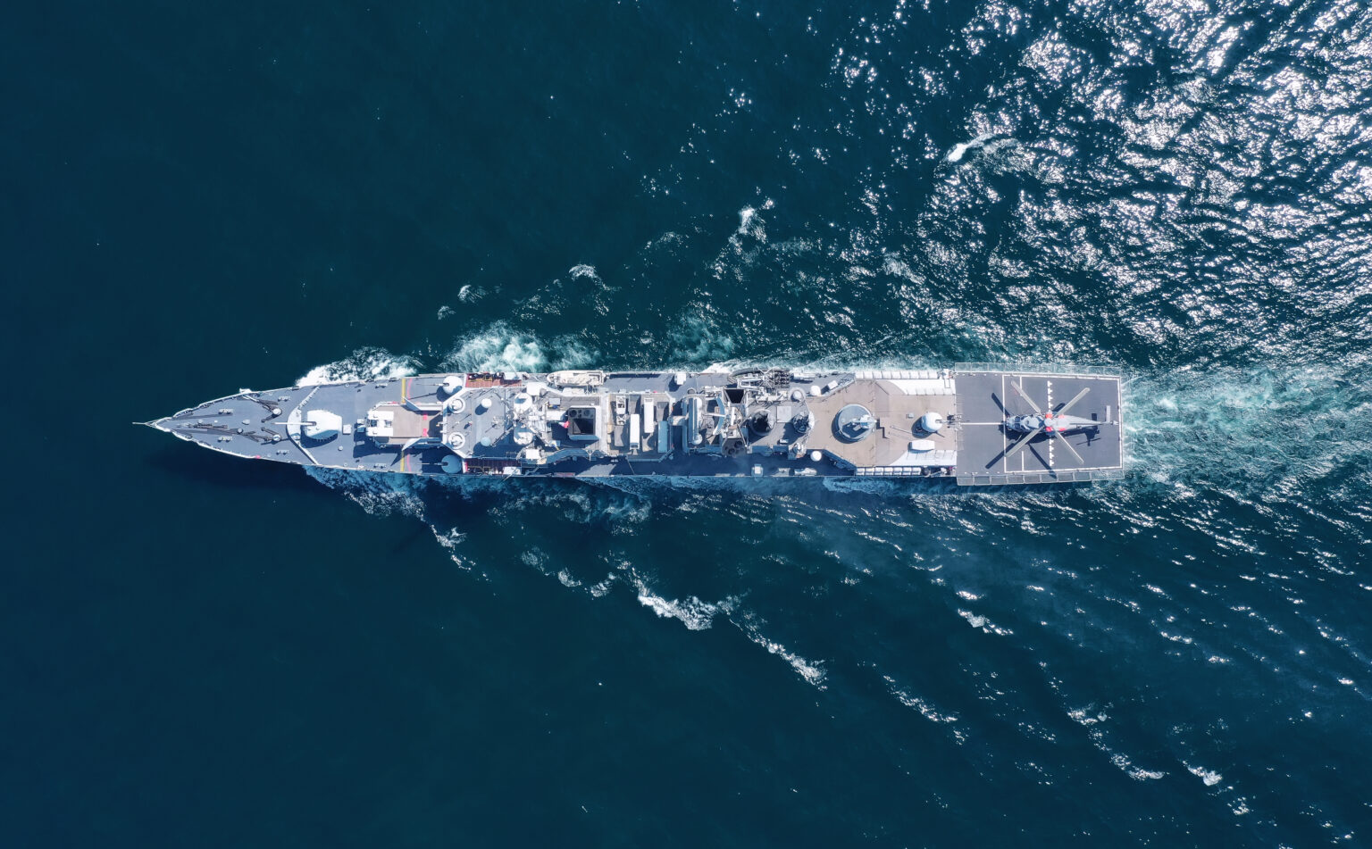 Aerial view of a naval warship with a helicopter on its helipad, sailing through the ocean.