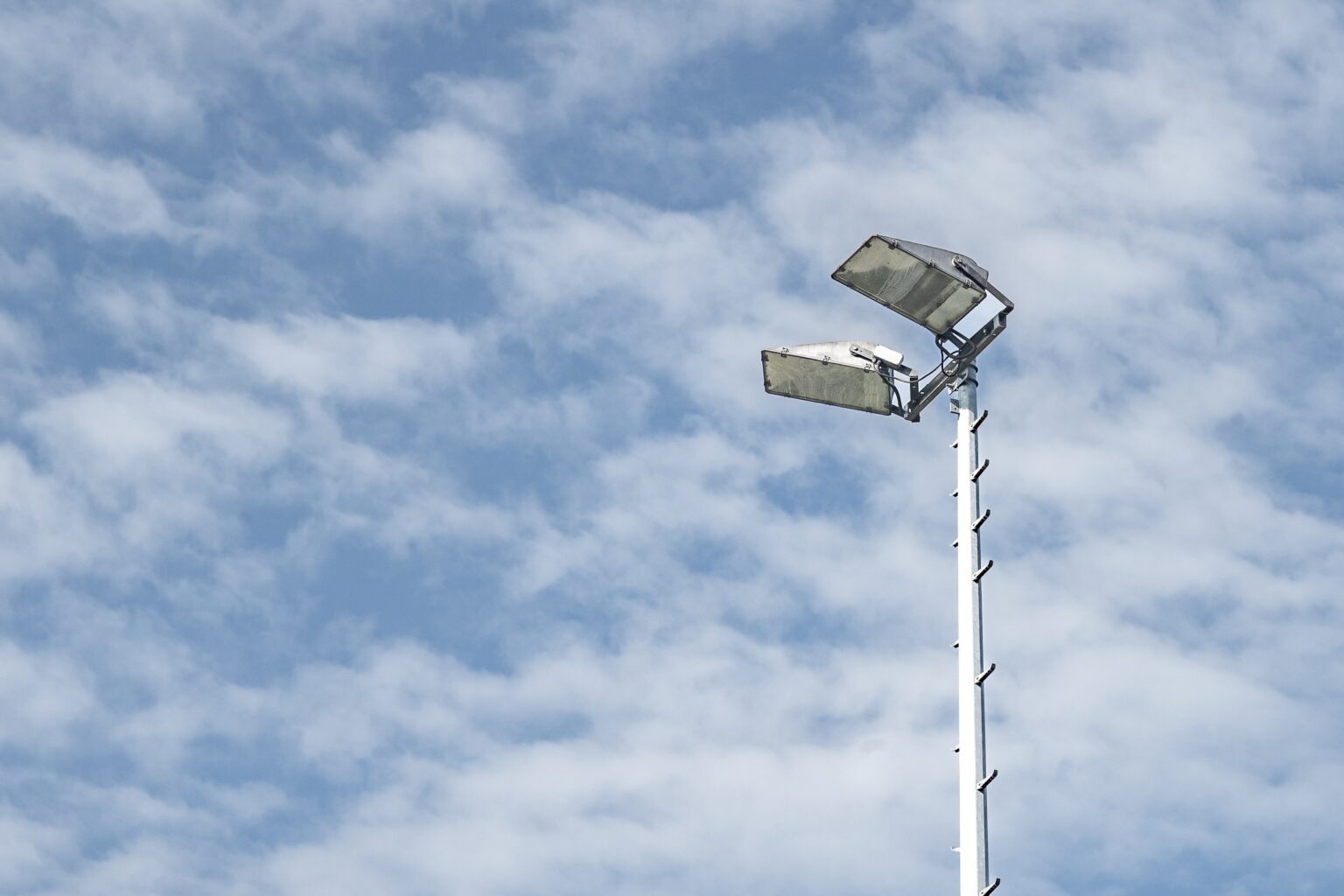 A tall floodlight pole with two large, slightly weathered lamps set against a bright blue sky with scattered white clouds